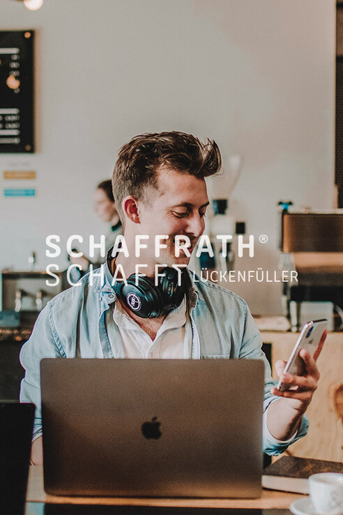 Platzhalterbild - Junger Mann der vor einem Laptop sitzt und ein Handy in der Hand hält